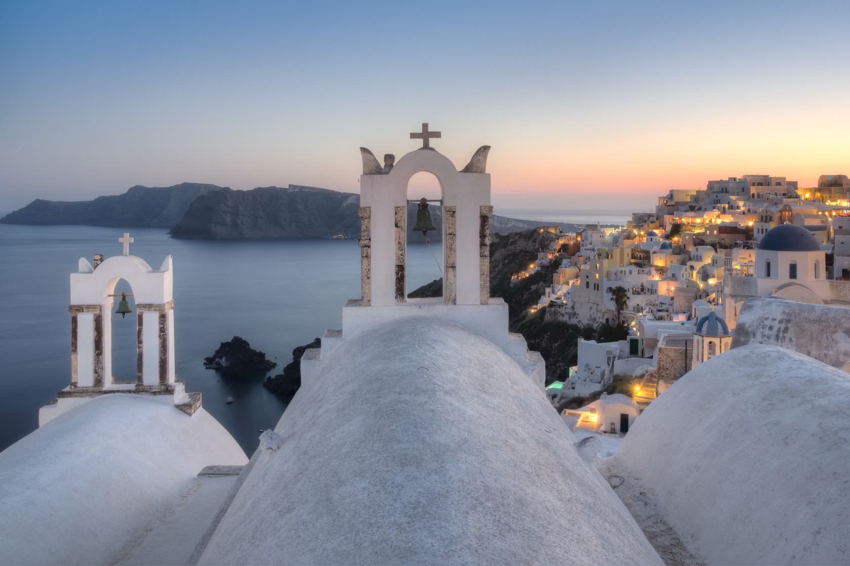 Oia at dusk on Santorini: two white bell towers above the caldera, the village lights coming on
